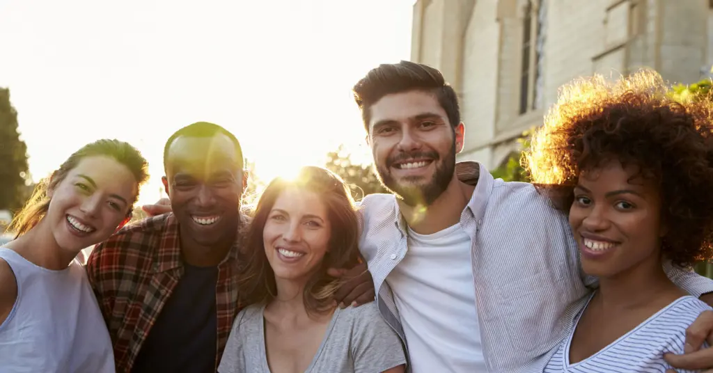 Five diverse adults hold each each shoulder-to-shoulder and smile, the sun shining bright behind them